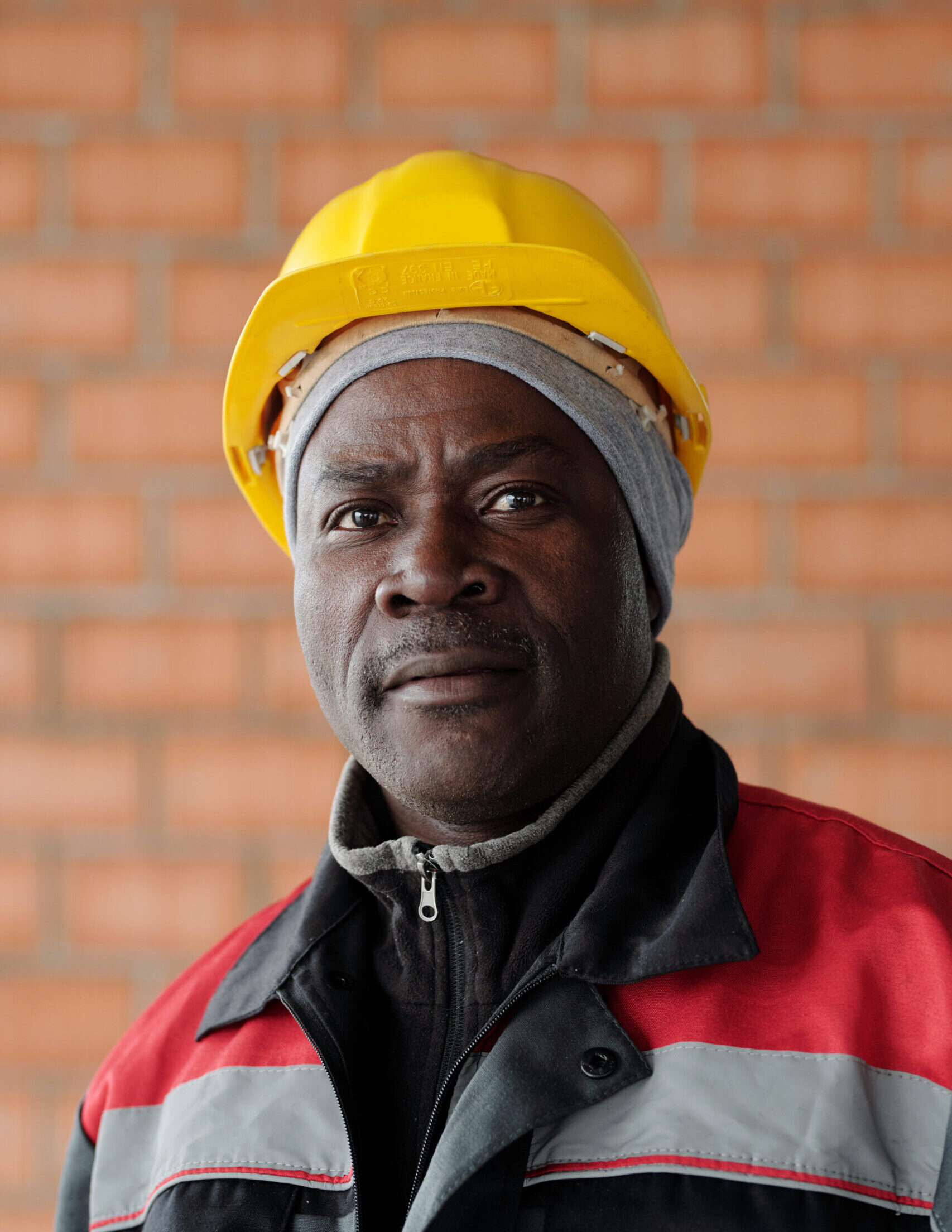 Modern experienced engineer or foreman in protective helmet looking at you while standing against brick wall at construction site
