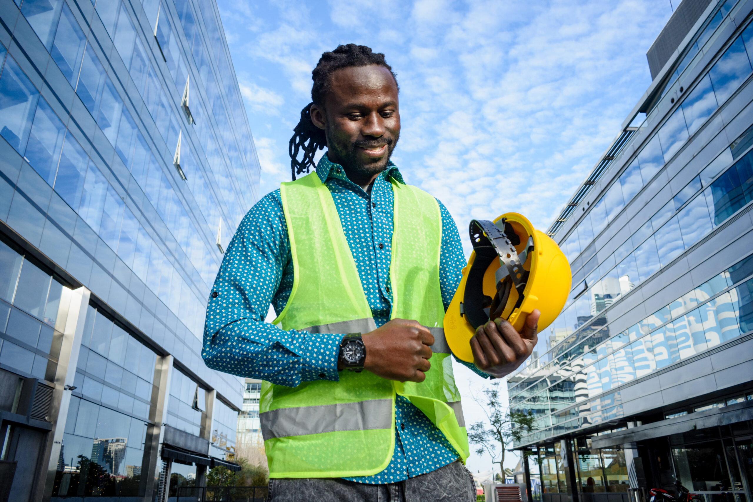 young civil engineer man of african ethnicity with beard and dreadlocks, holding his helmet with hand is standing outdoors smiling happy at end of work day